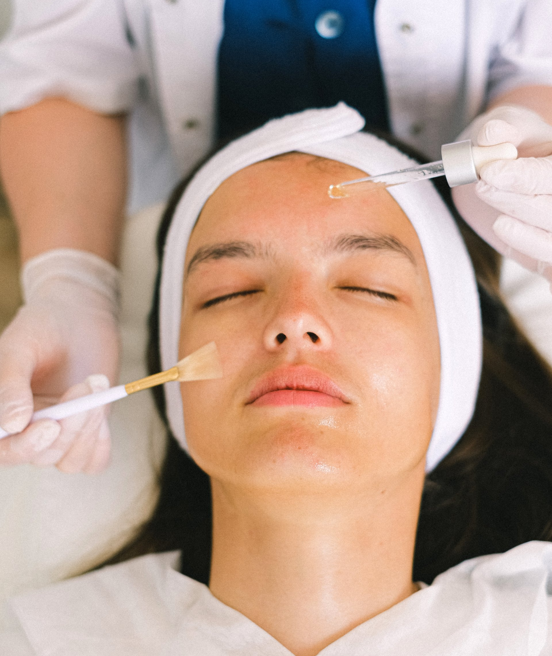 Woman getting facial peel at a spa, eyes closed. Aesthetician applies product with a brush and dropper.