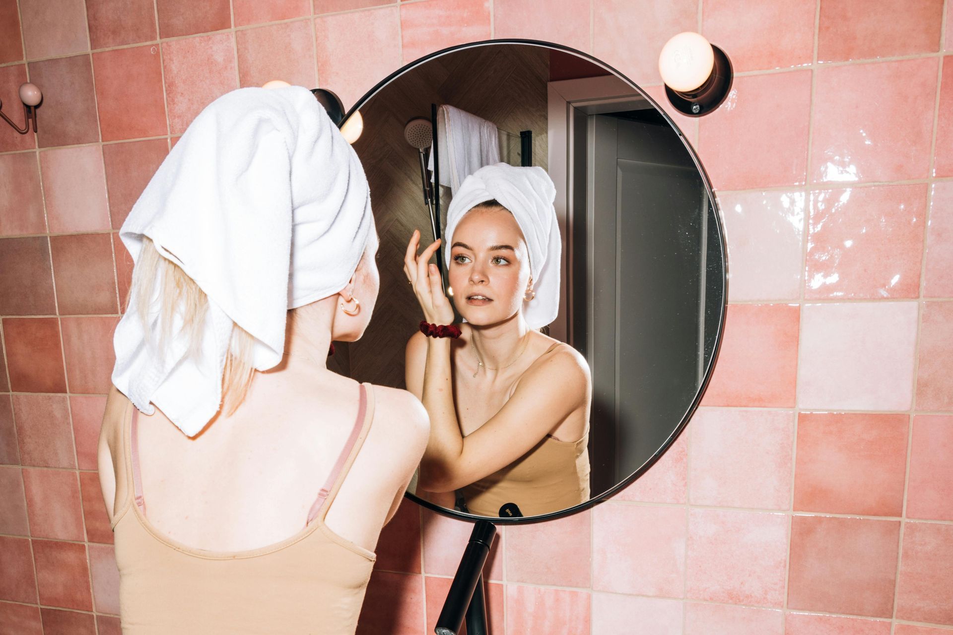 Een vrouw in een beige top en handdoek kijkt naar haar droge huid in een ronde spiegel.