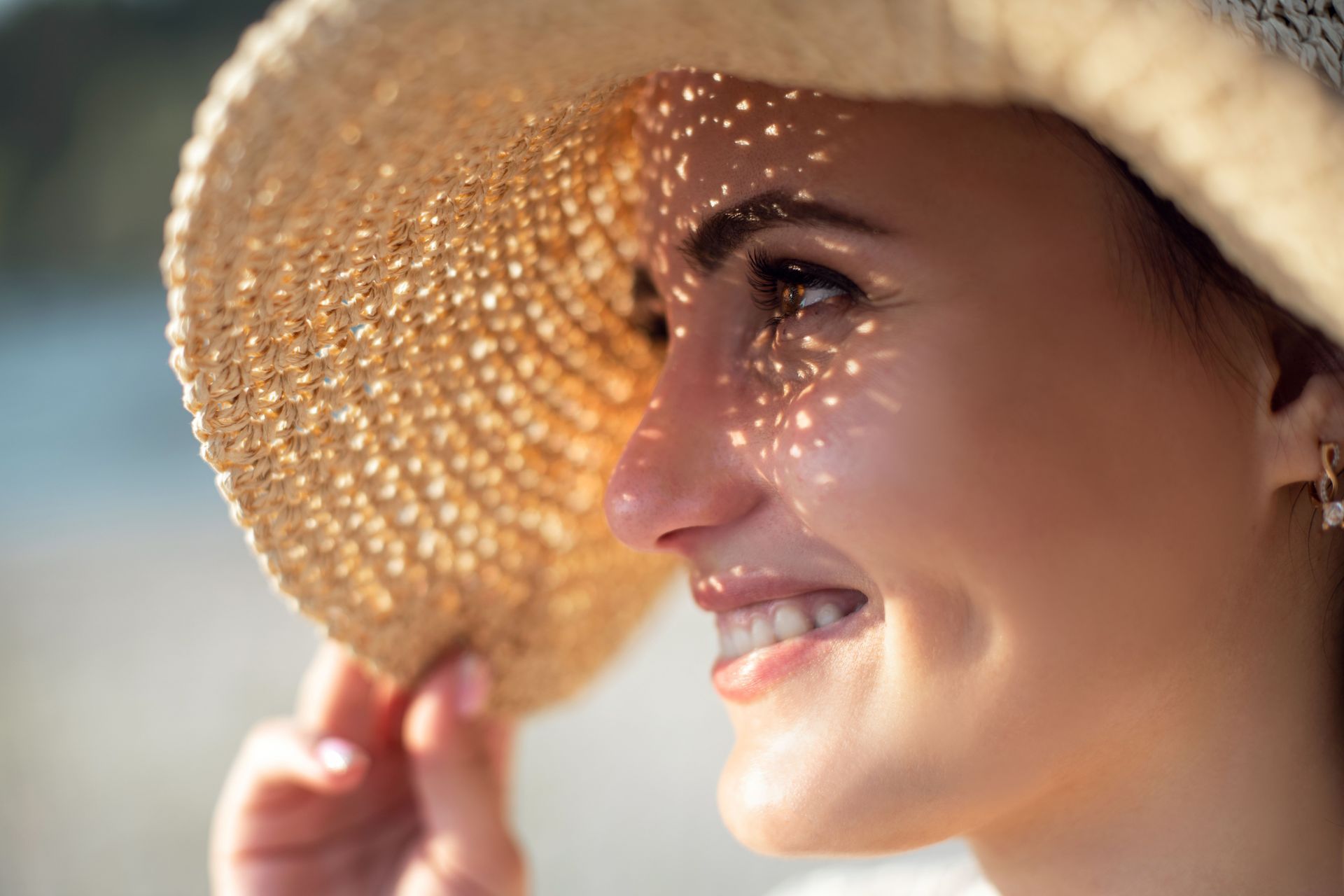Smiling woman in a straw hat at the beach, holding the brim in bright sunlight