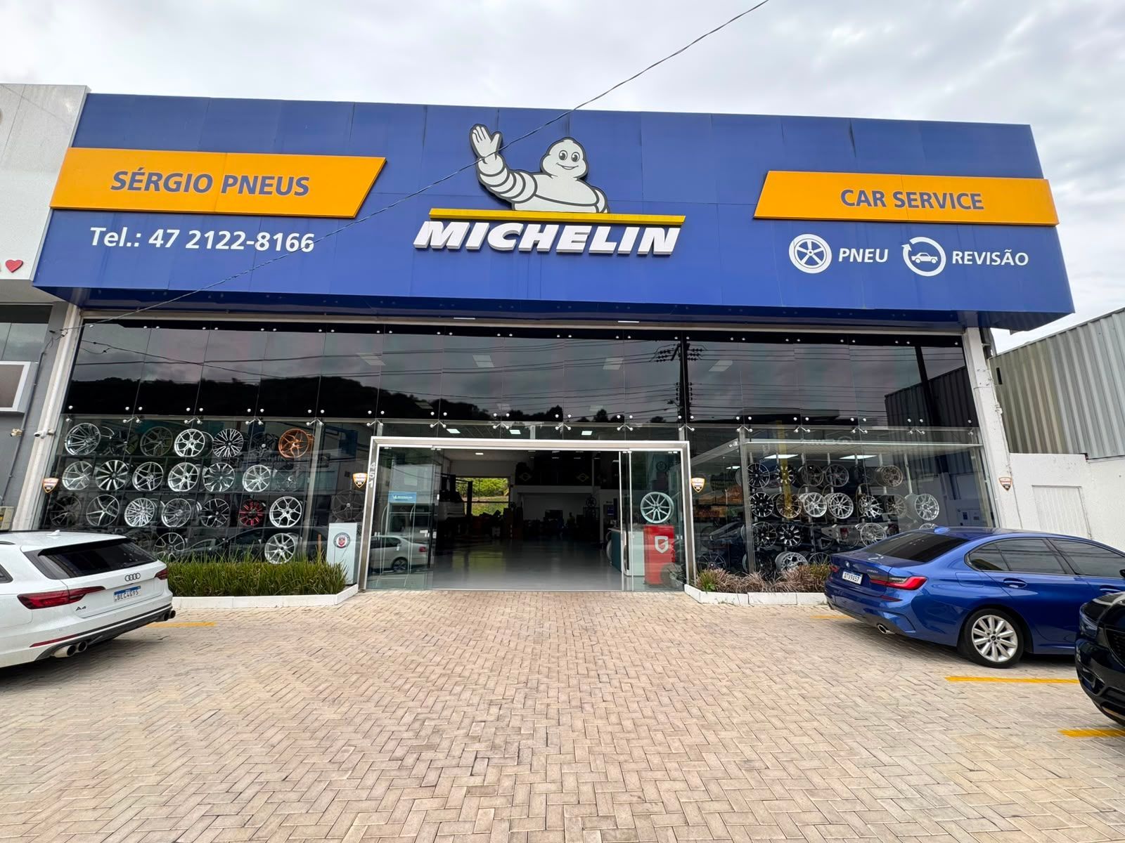 Fachada da loja de pneus Michelin com sinalização azul e laranja, carros estacionados e entrada de vidro.