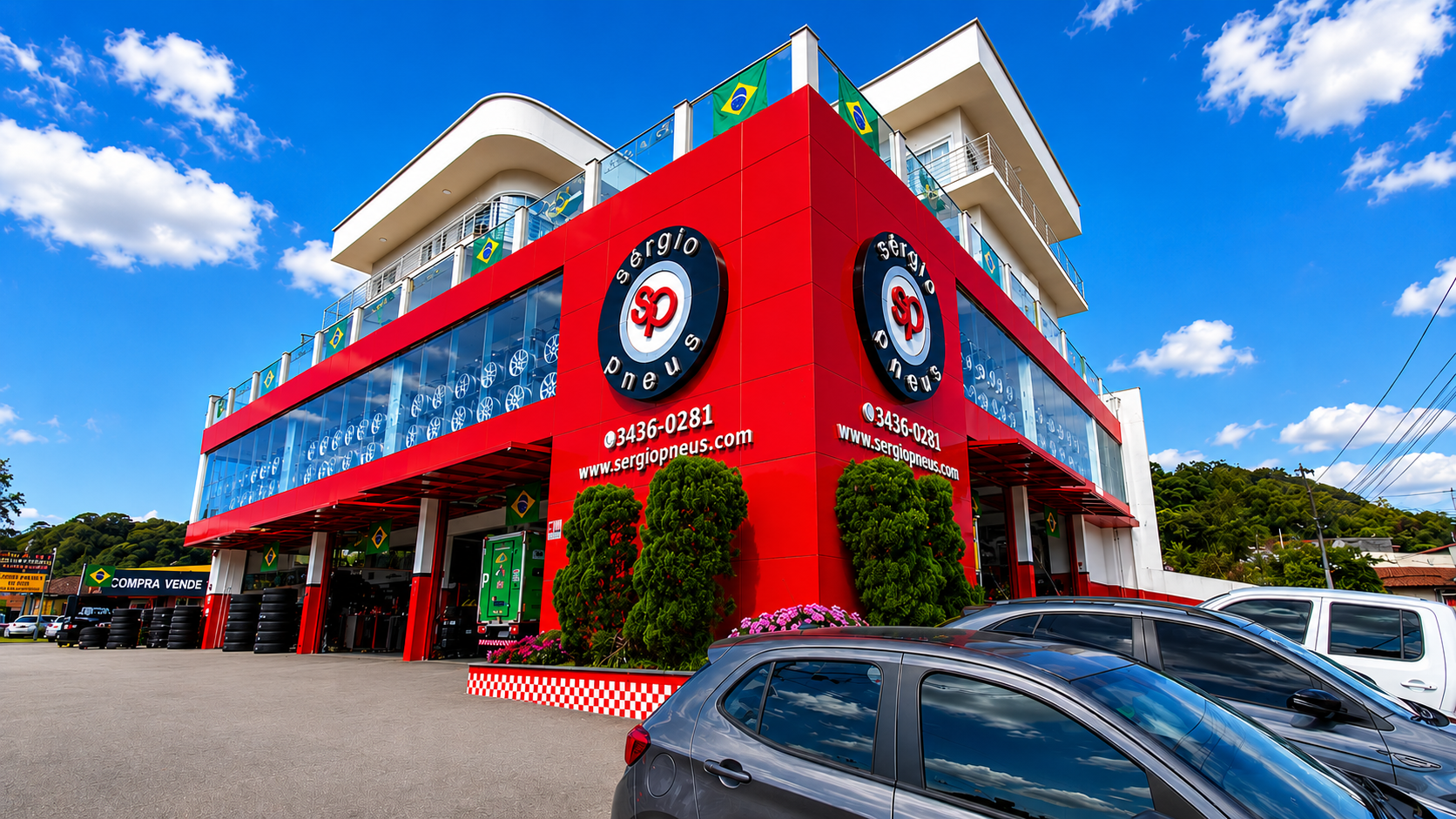 Prédio vermelho com grandes logotipos circulares, janelas azuis e carros estacionados sob um céu azul brilhante.