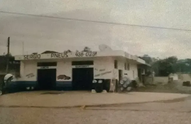 Pequeno prédio branco à beira da estrada, com portas azuis e várias motocicletas estacionadas em uma esquina empoeirada.
