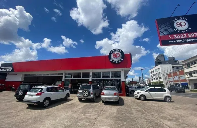 Concessionária de veículos com toldo vermelho, carros estacionados e uma grande placa à beira da estrada sob um céu azul.