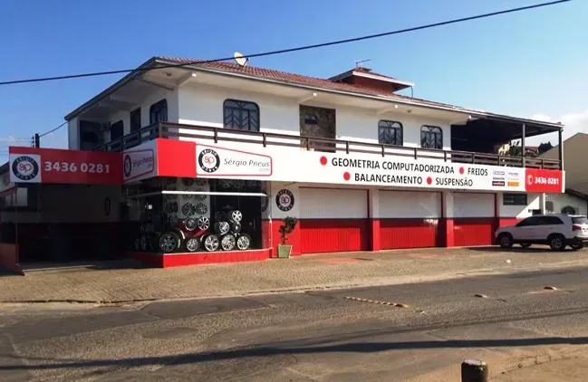 Prédio comercial de dois andares com sinalização vermelha e branca em uma esquina ensolarada.
