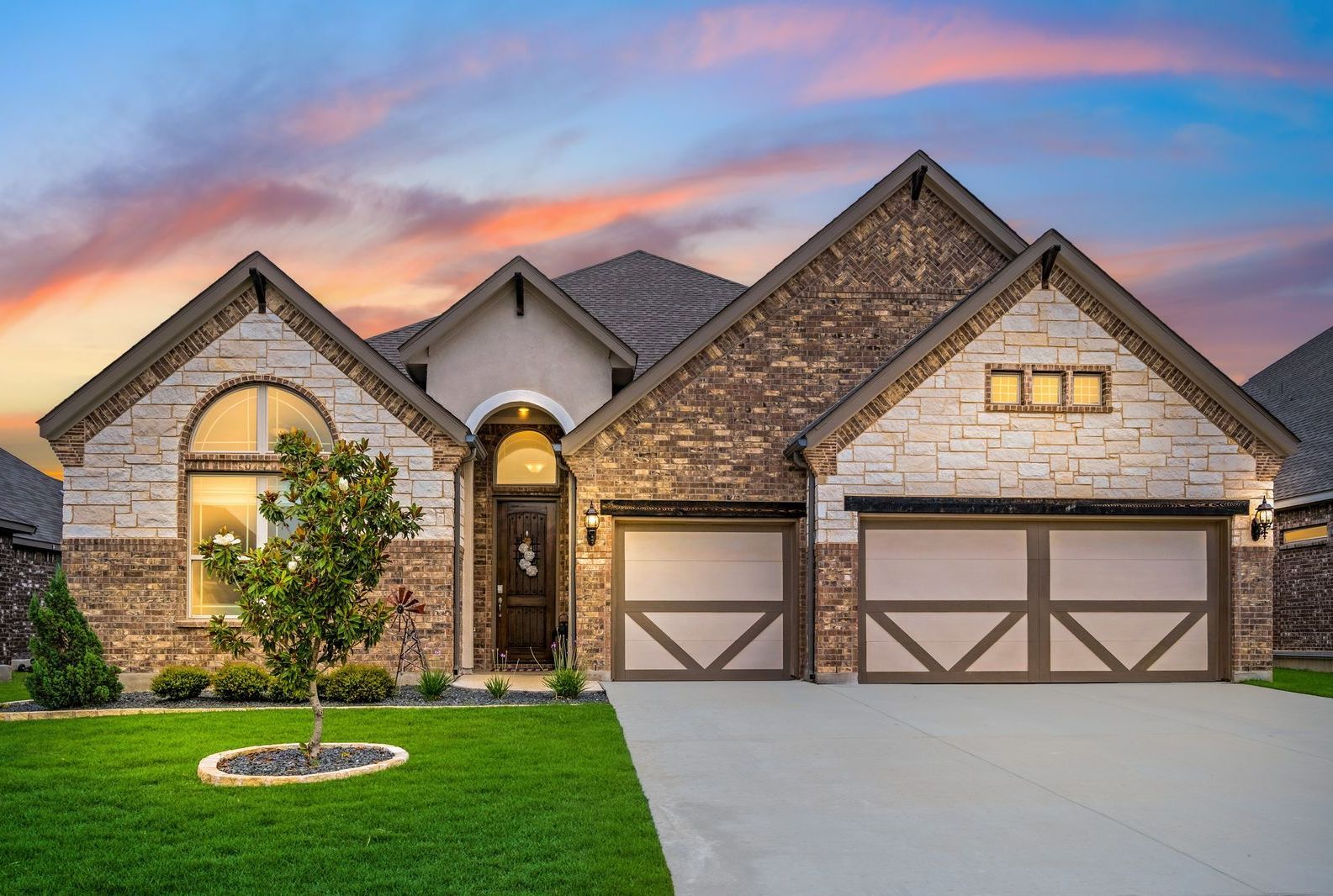 A large brick house with two garage doors and a tree in front of it.