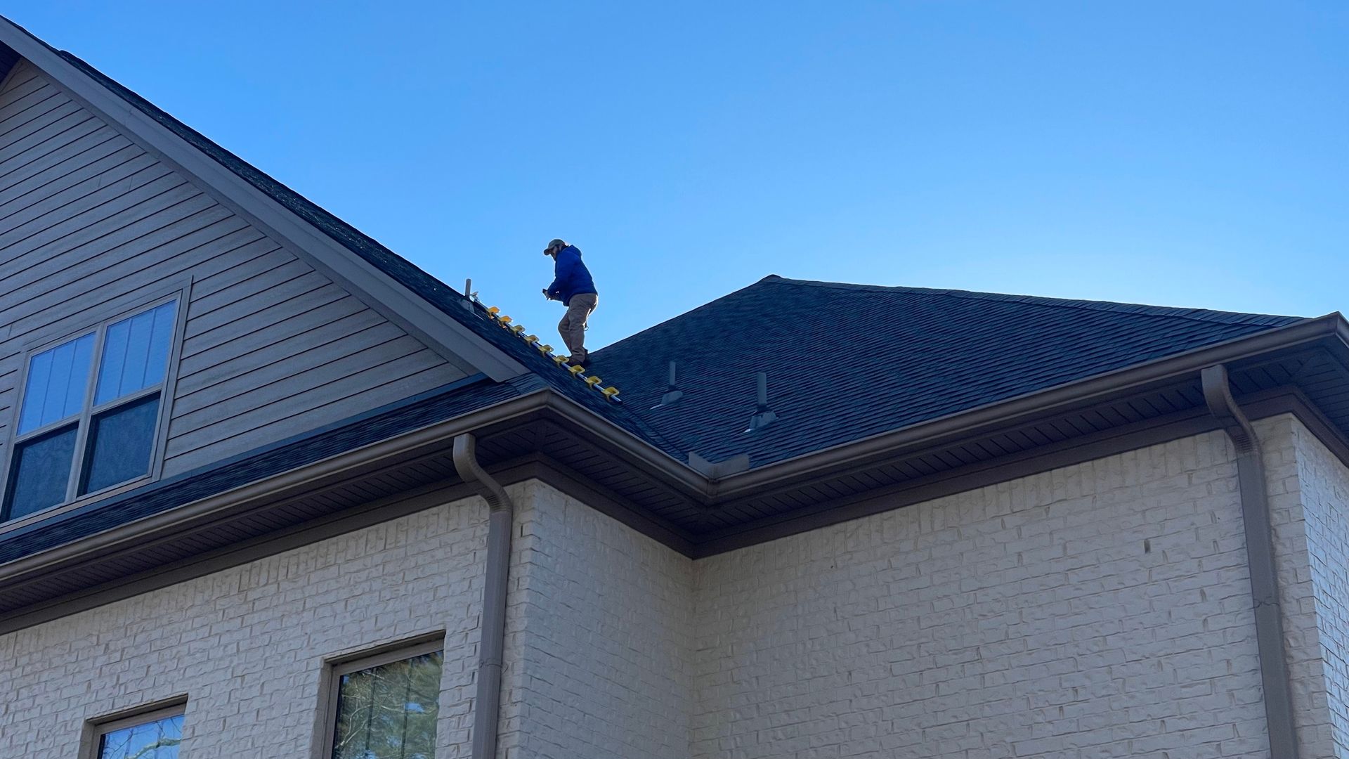 Person on a roof, near the edge, working on a dark roof with gutters, a clear blue sky.