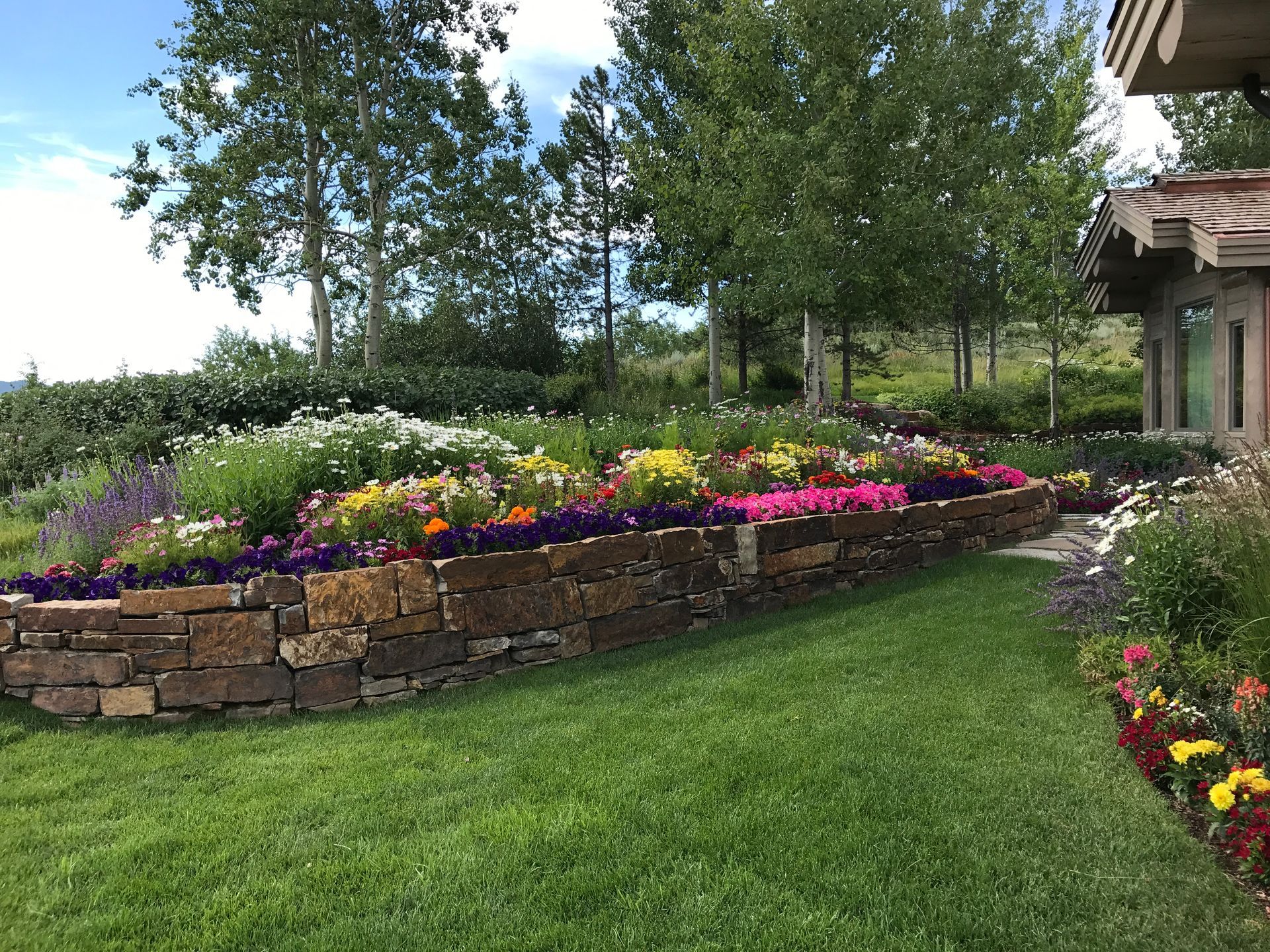 Colorful flower beds and a lush green lawn beside a stone retaining wall on a sunny garden hillside.