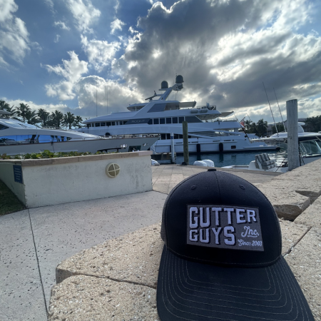 Black Gutter Guys cap on a dock with a luxury yacht and cloudy sky in the background