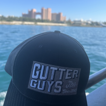 Black “Gutter Guys” cap in the foreground, with water and a blurred city skyline in the background.
