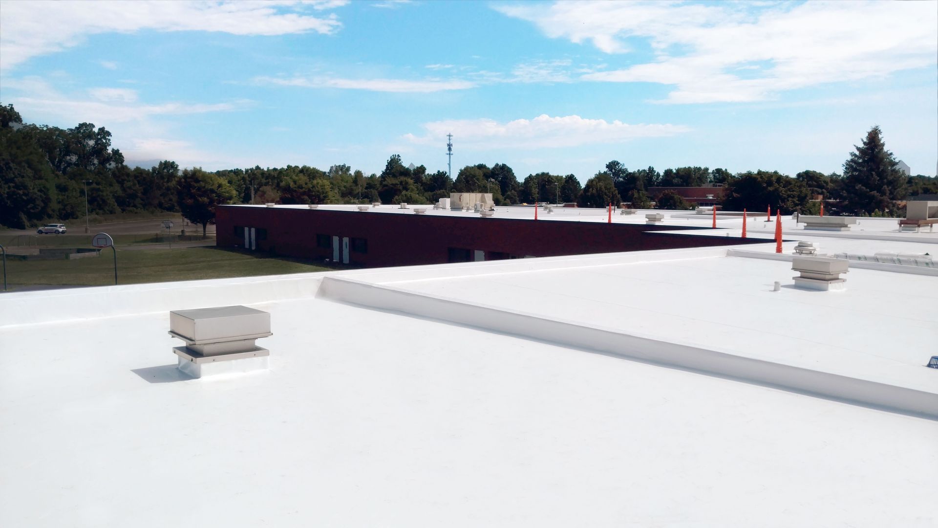 Flat white rooftop with vents and red skylights, overlooking trees and low buildings under a clear sky