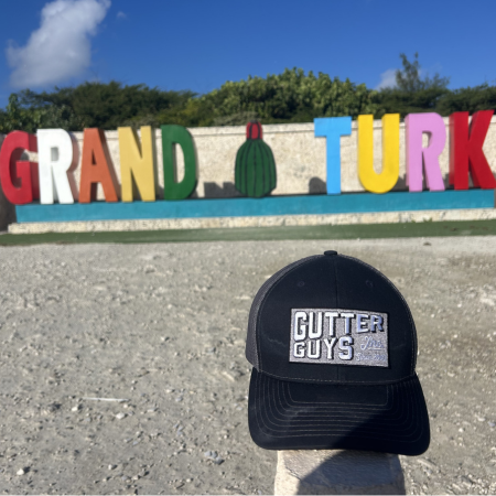 Black Gutter Guys cap in foreground with colorful Grand Turk sign on a sunny beach backdrop