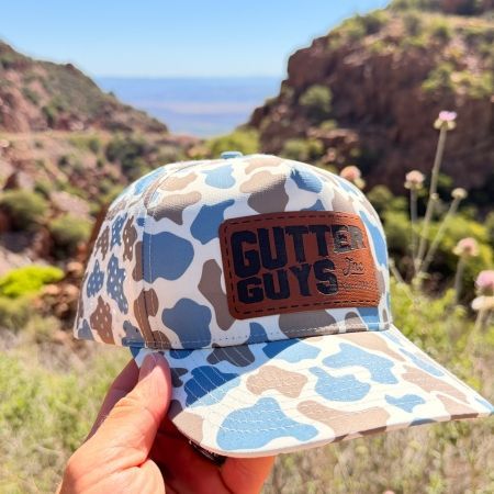 Black “Gutter Guys” cap and compass with “Stay Tuned Coming Soon” text on torn paper background