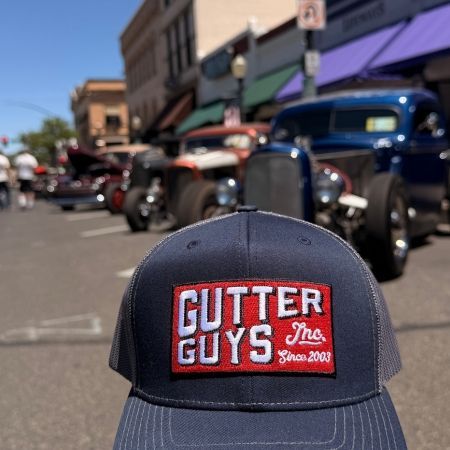 Black “Gutter Guys” cap in the foreground, with water and a blurred city skyline in the background.