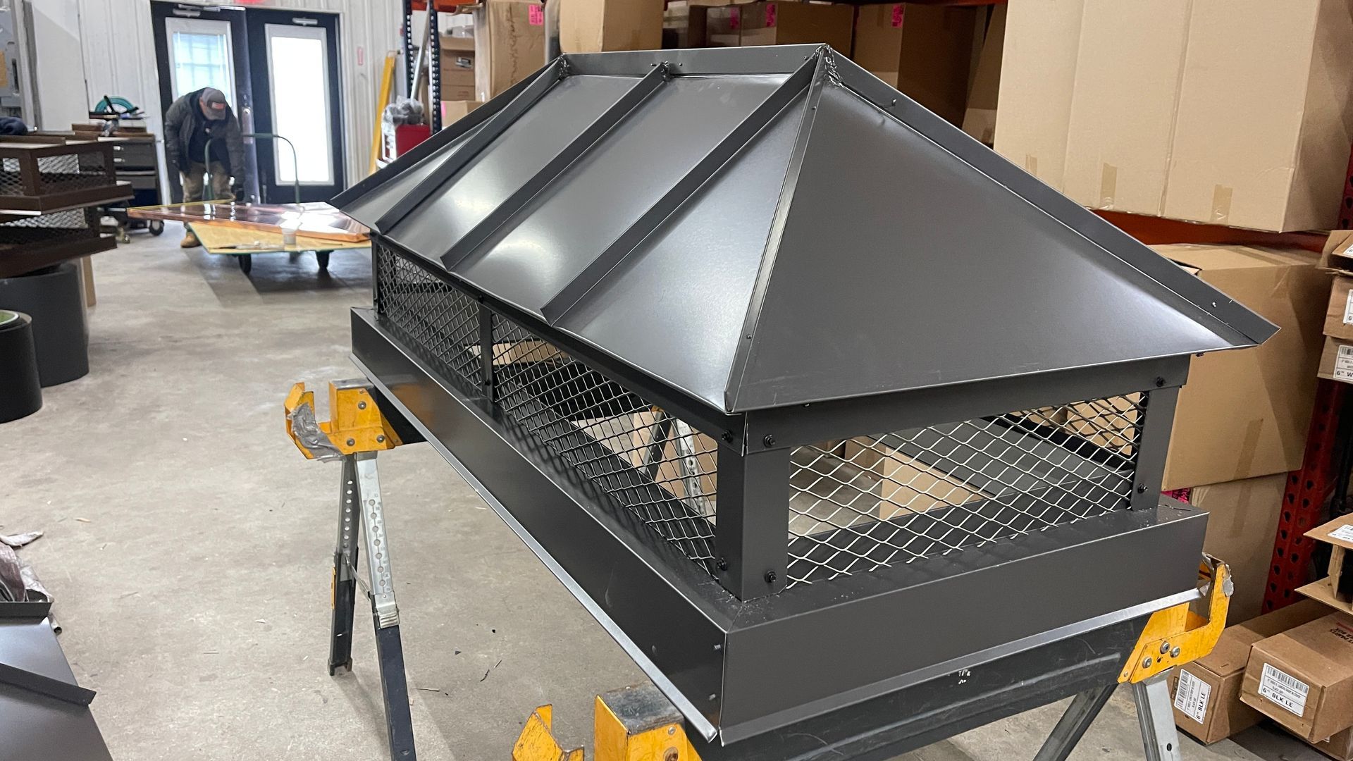 Black metal rooftop vent or chimney cap in a workshop, with conical top and mesh sides on a cart