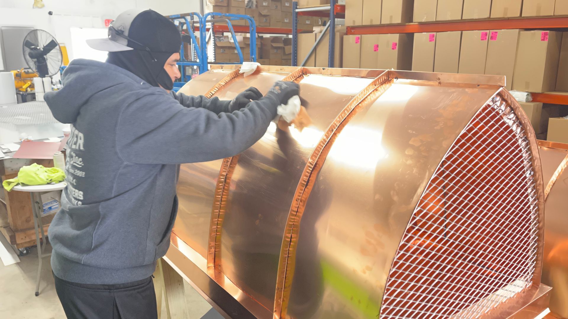 Worker in a gray hoodie welds a large copper cylinder in an industrial workshop.