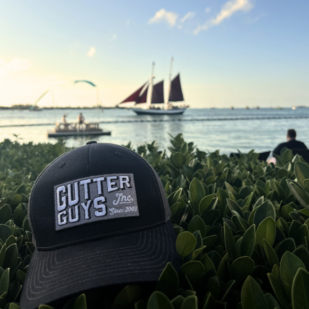Black “Cutter Guys” cap in foreground with sailboats and people on the water at sunset