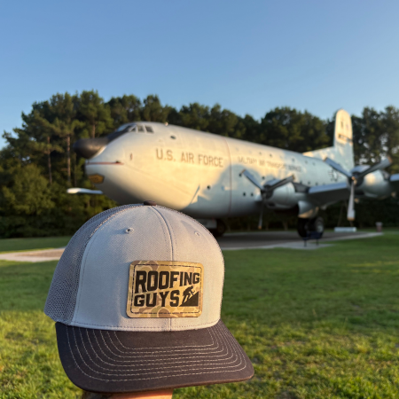 Black “Gutter Guys” cap in the foreground, with water and a blurred city skyline in the background.