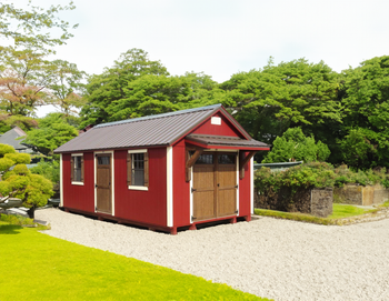 Large red barn with white trim, metal roof, wooden doors, windows, covered porch on gravel drive in landscaped yard.