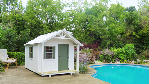 Small white storage shed with peaked roof, green trim, and front porch beside turquoise swimming pool in landscaped backyard.