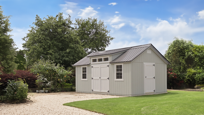 Large tan storage shed with charcoal metal roof, dormer windows, transom lights, white double doors on gravel pad in backyard