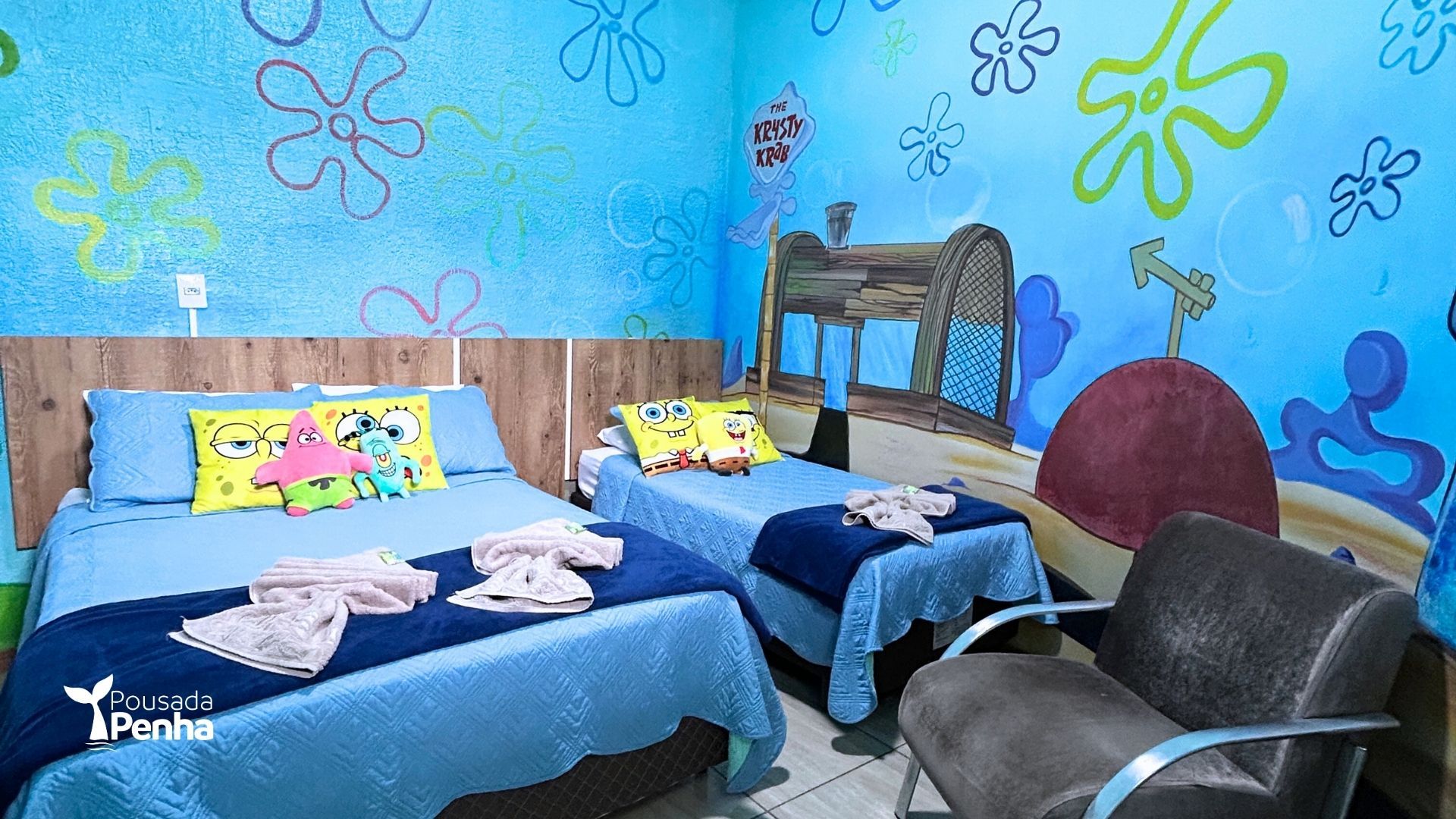 Duas camas azuis em um quarto colorido com murais florais nas paredes e uma cadeira preta junto à janela.