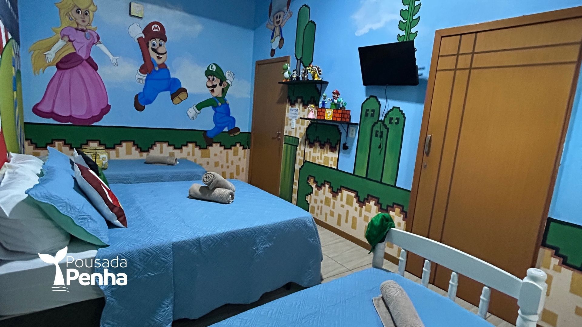 Quarto temático do Mario com paredes azuis, duas camas de solteiro, personagens em um mural e uma TV fixada na parede.