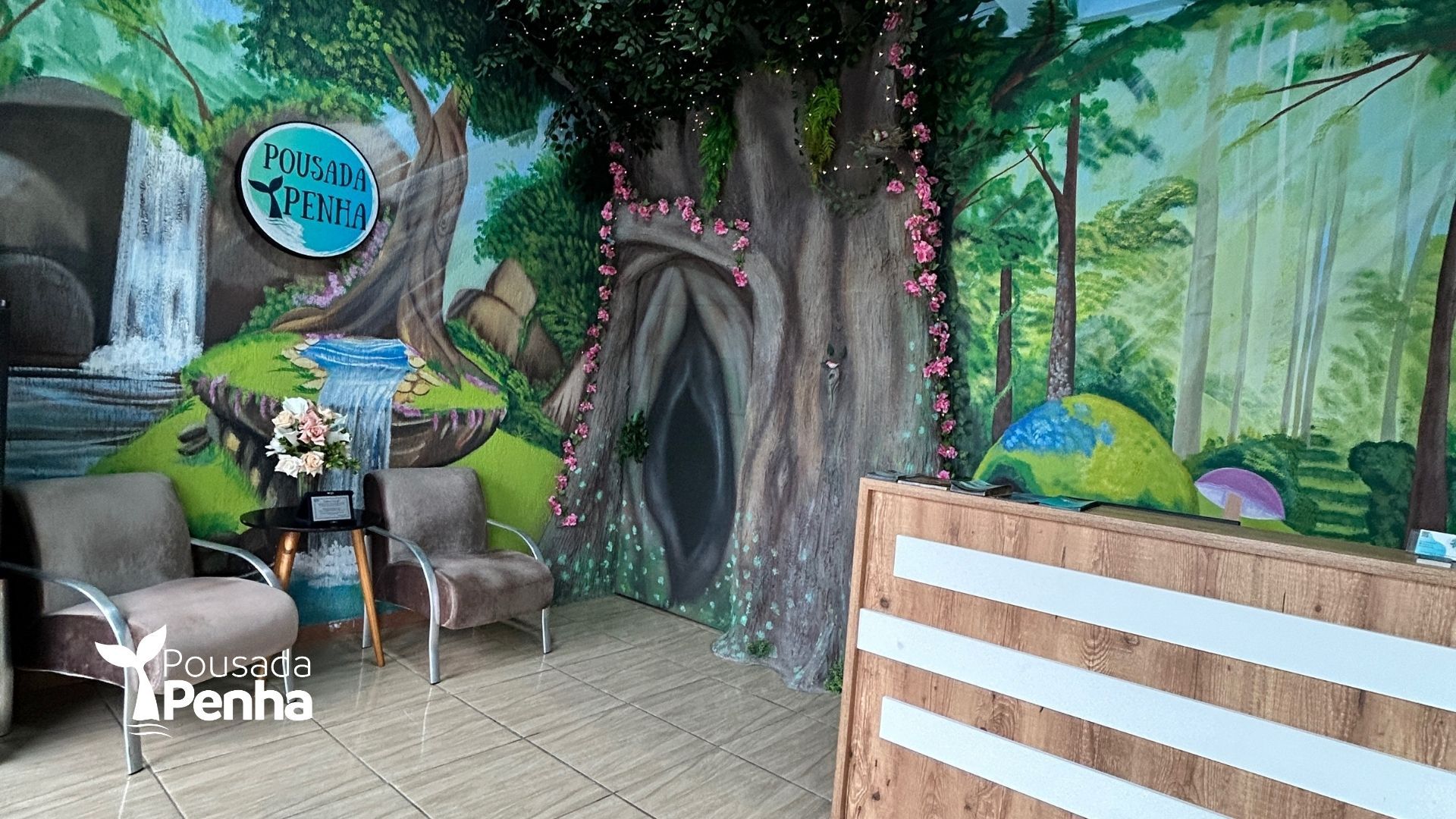 Pessoa sentada em uma área com temática de floresta, ao lado de uma pequena mesa e um banco, com um grande mural de árvore atrás dela.