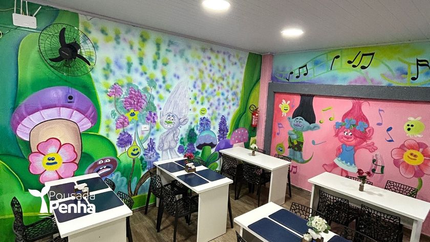 Mural colorido na sala de aula com flores, borboletas e personagens de desenho animado, além de carteiras em primeiro plano.