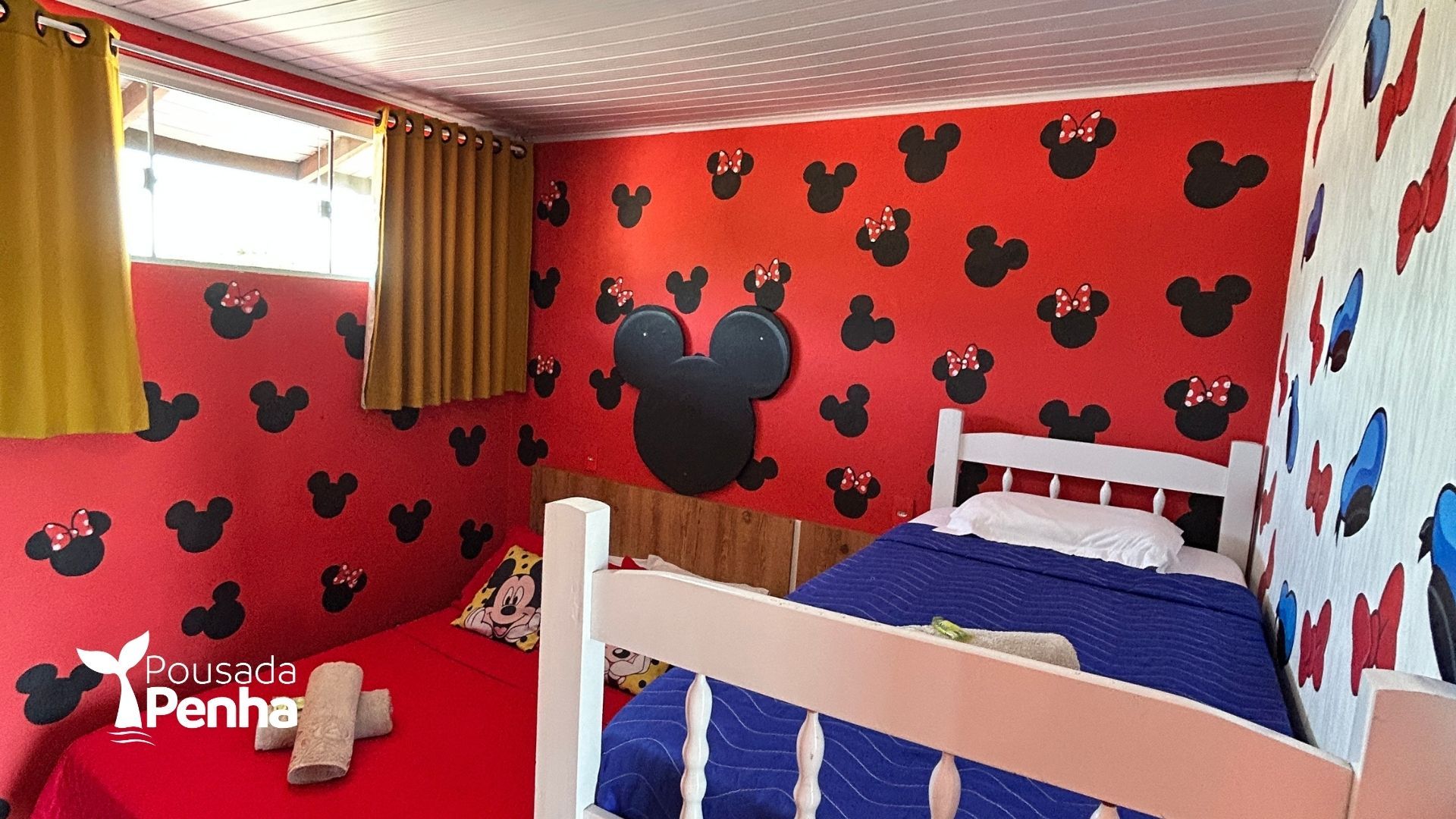 Quarto temático do Mickey Mouse com paredes vermelhas, cama azul e beliches brancos perto de uma janela.