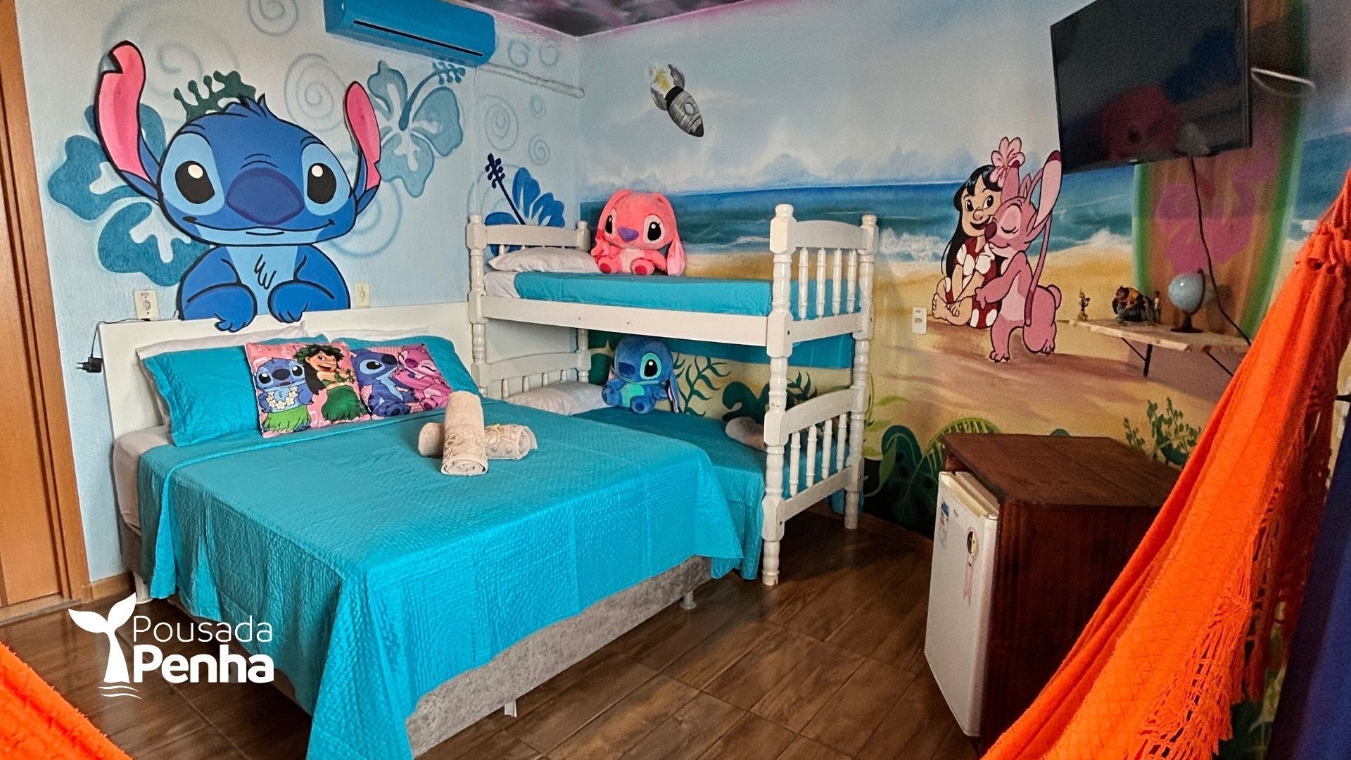 Quarto infantil colorido com duas camas, decoração de parede com desenhos animados, roupa de cama turquesa e uma cadeira laranja em formato de tenda.