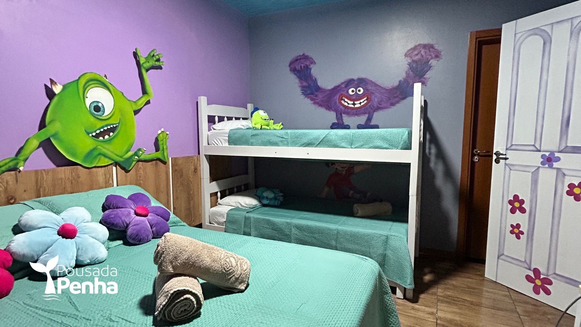 Quarto infantil colorido com beliche, roupa de cama azul-petróleo, bichos de pelúcia e adesivos de parede com monstros de desenho animado.
