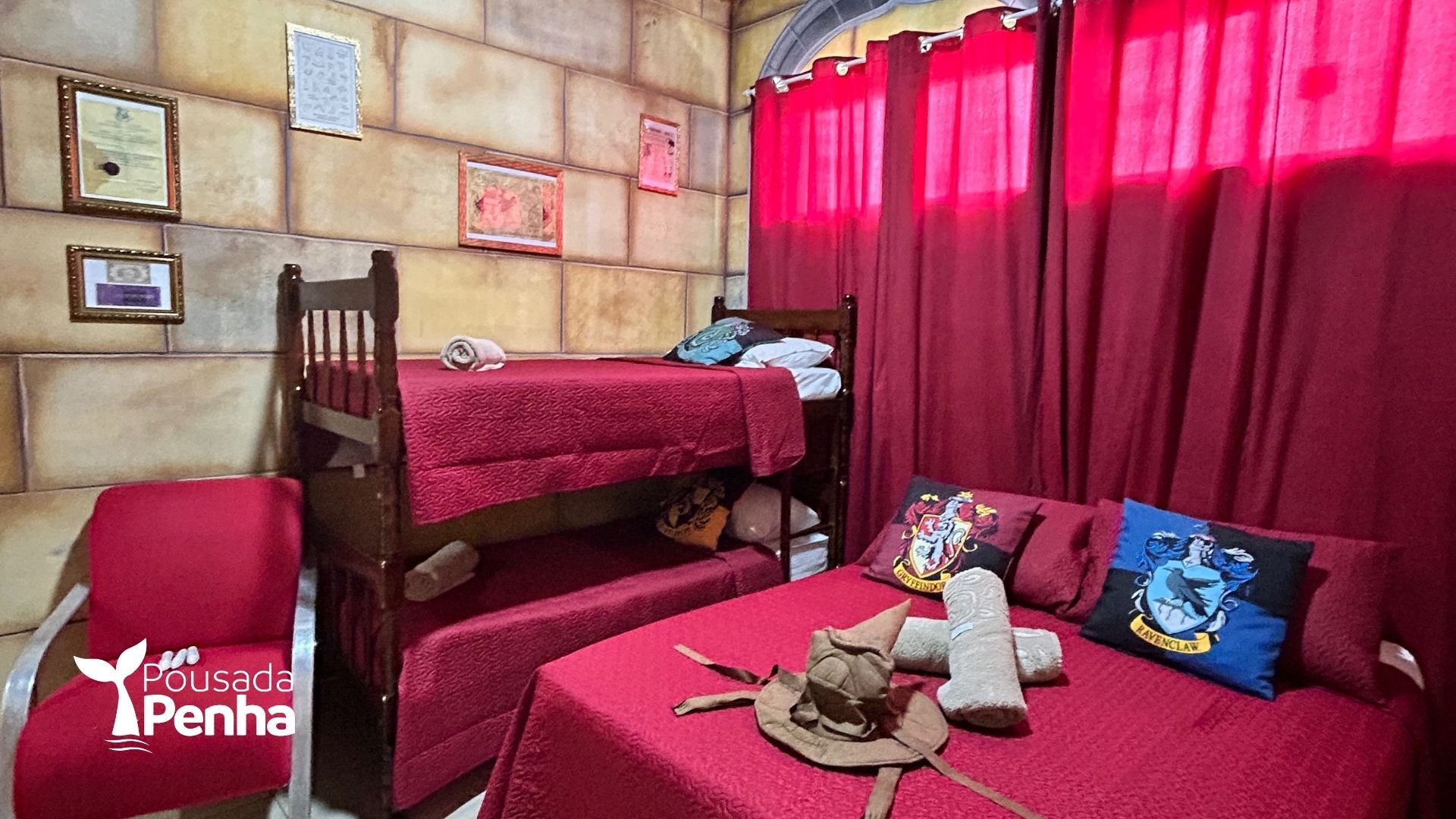 Quarto pequeno com cortinas vermelhas, duas camas e uma mesa com bichos de pelúcia e almofadas.