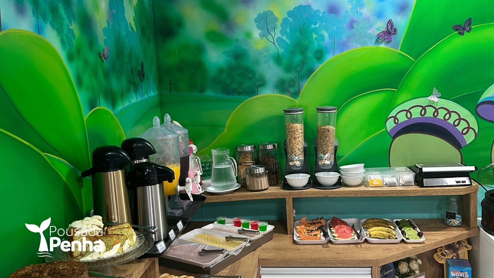Mesa de buffet com café, doces e frutas em frente a um mural verde vibrante.