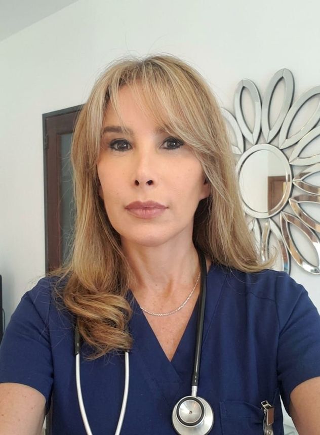 Mujer con blusa azul y estetoscopio, de pie en el interior de un edificio, apoyada contra una pared clara.