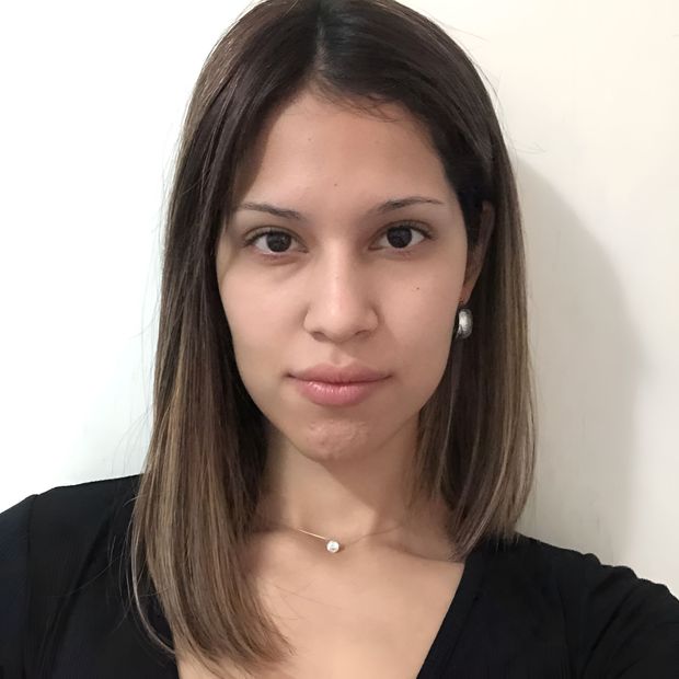 Retrato de una mujer con cabello castaño hasta los hombros, vestida con una blusa negra y un collar, frente a una pared lisa.