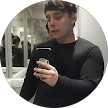 Una persona con una camisa negra se toma una selfie frente al espejo en un baño, sosteniendo un teléfono y mirando hacia abajo.