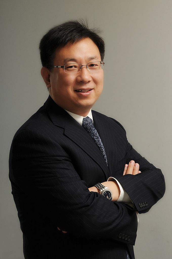 Woman with short black hair, wearing a white shirt, navy cardigan, and hands in pockets, smiling against a blue background.