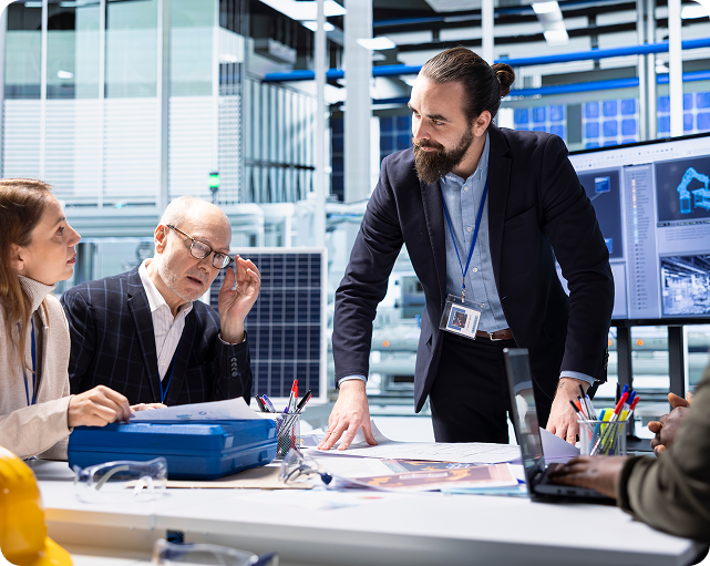 People at a table, collaborating on blueprints in an office with a large screen displaying schematics.