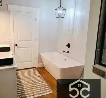 Bathroom with white tub, door, and walls. Brown wooden floor and small rug. Black light and faucet.