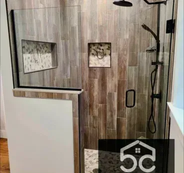 Shower with wood-look tile, glass door, and black fixtures. Features recessed storage and contrasting tile accents.