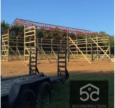 Construction site: Wooden frame of a large building with a red steel roof. A trailer is in the foreground.