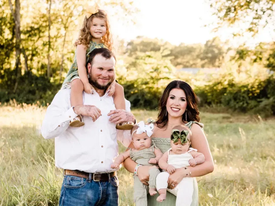 Family portrait in a field: father with daughter on shoulders, mother with twin babies. All smiling. Golden sunlight.