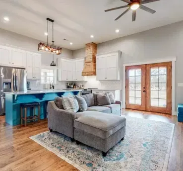 Open-concept living space with kitchen and living room. Teal island, gray couch, wooden floors, French doors.