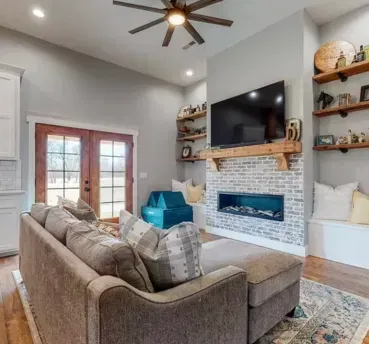Cozy living room with gray walls, fireplace, TV, and brown sectional sofa. Wooden shelves and French doors visible.