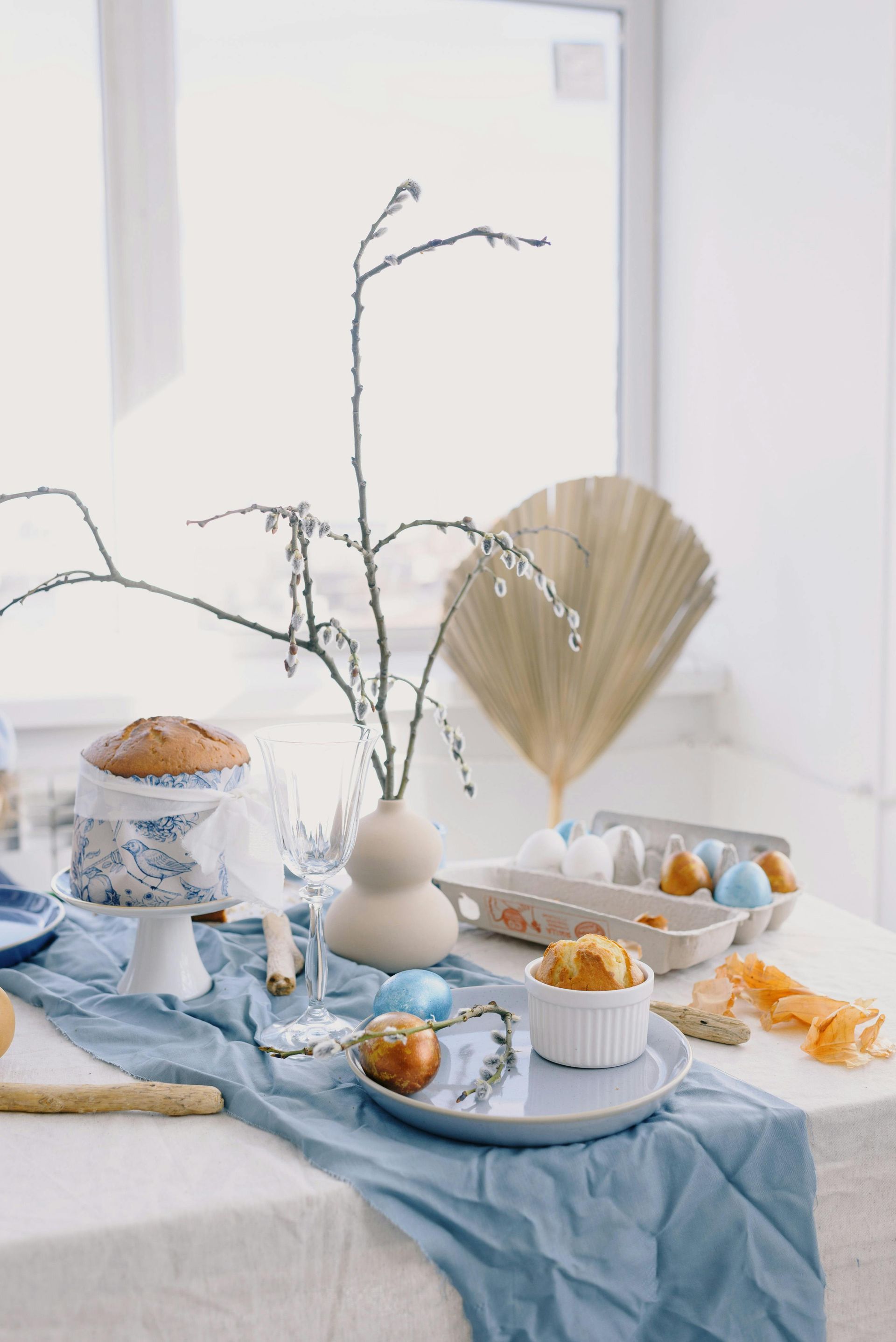 Easter table setting with cake, eggs, blue cloth, and floral decor near a window.