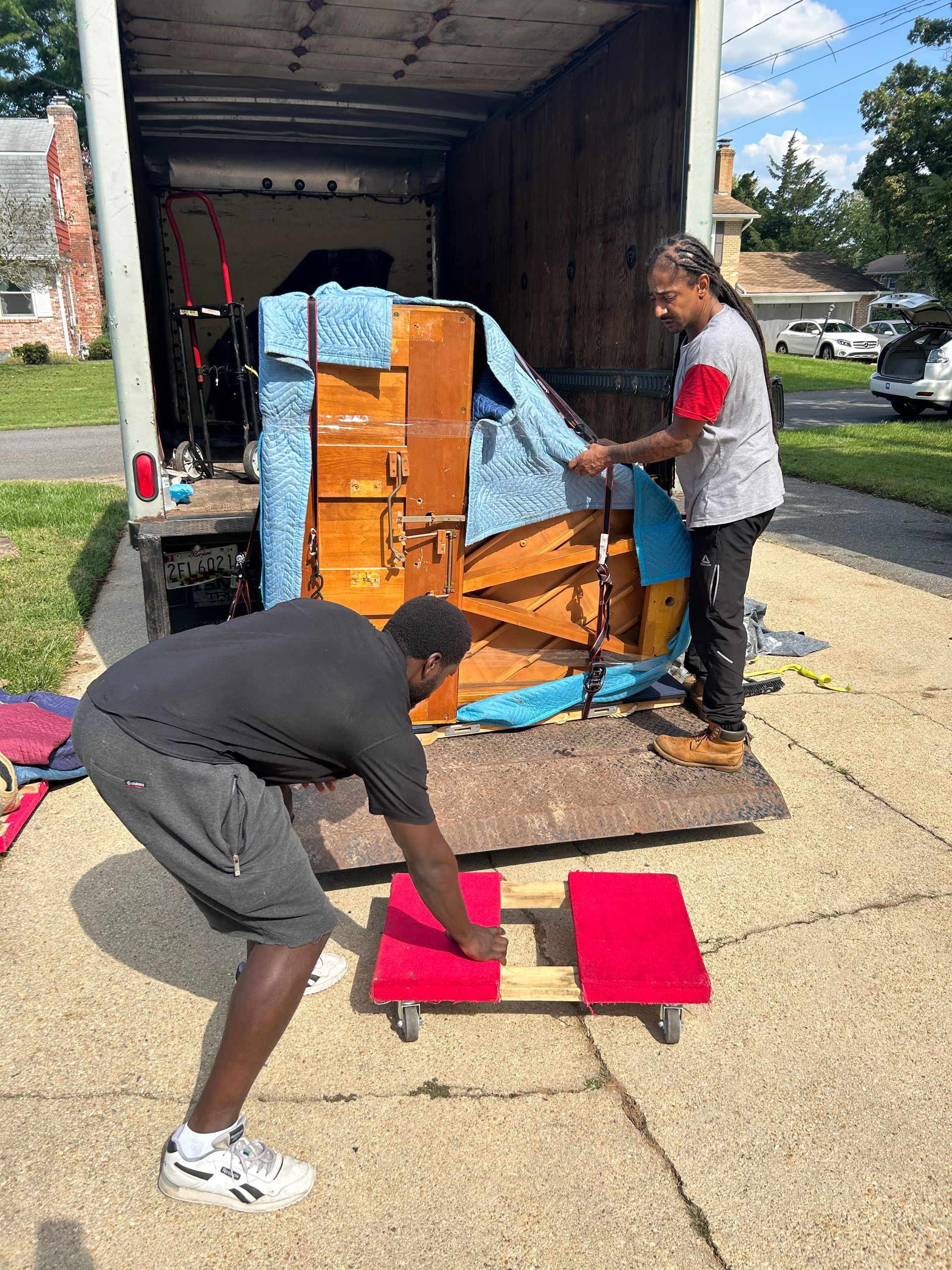 Two movers prepare to unload a large, blanket-wrapped piano from a moving truck onto a red wooden dolly in a driveway.