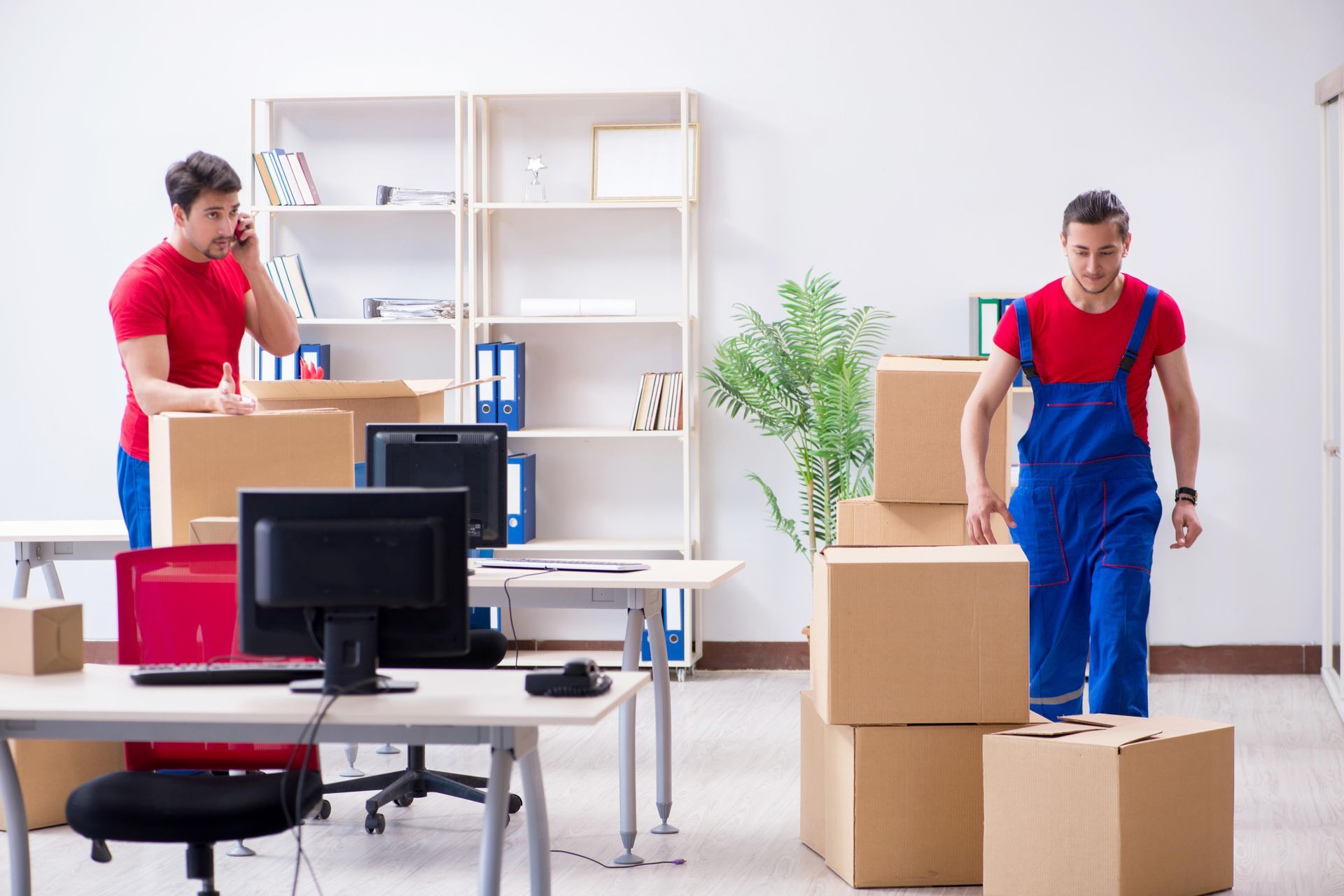 Two movers in red shirts and blue overalls carrying boxes in a bright office with desks and shelves