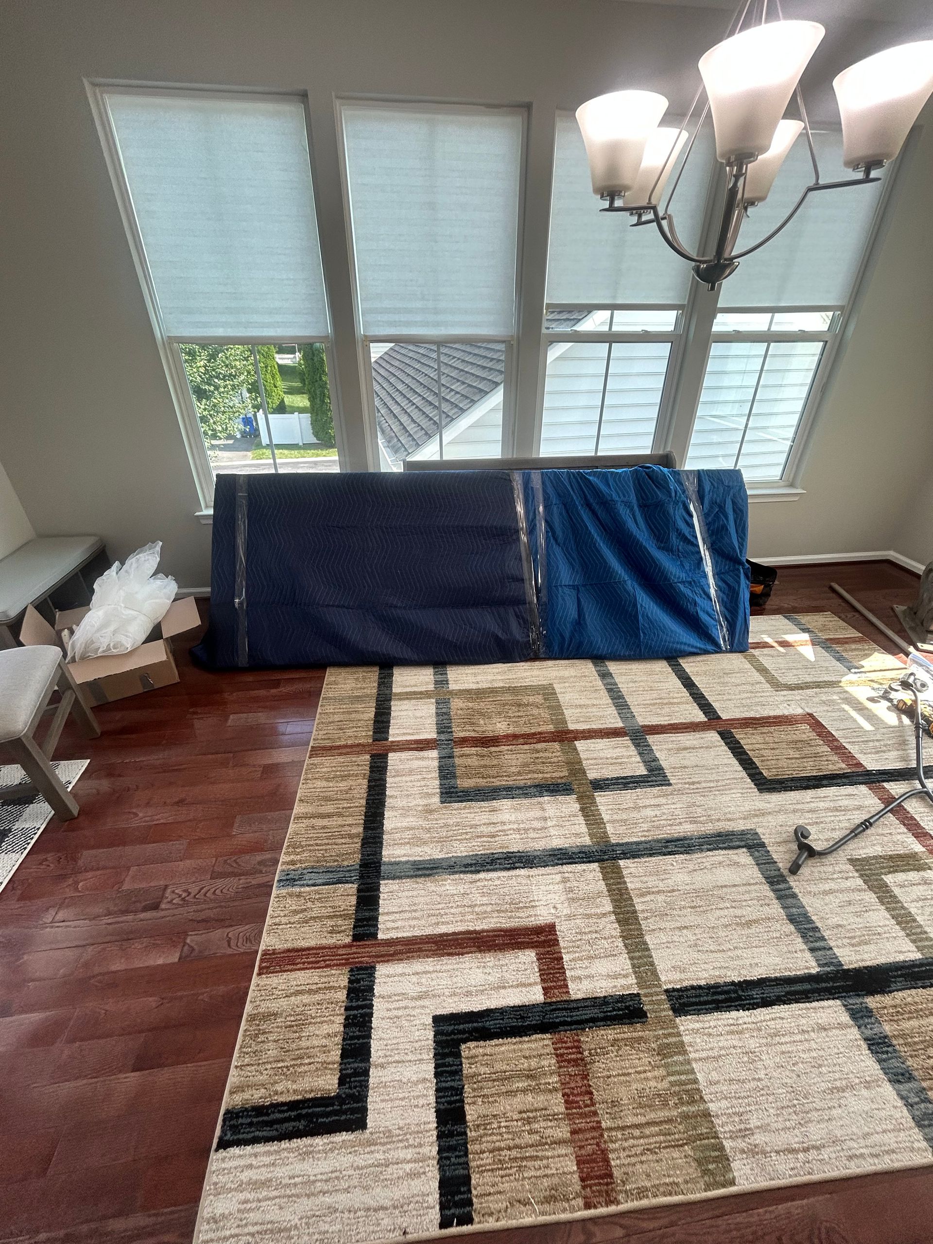 Two large items wrapped in blue moving blankets sit on a patterned rug in a dining room with windows and a chandelier.