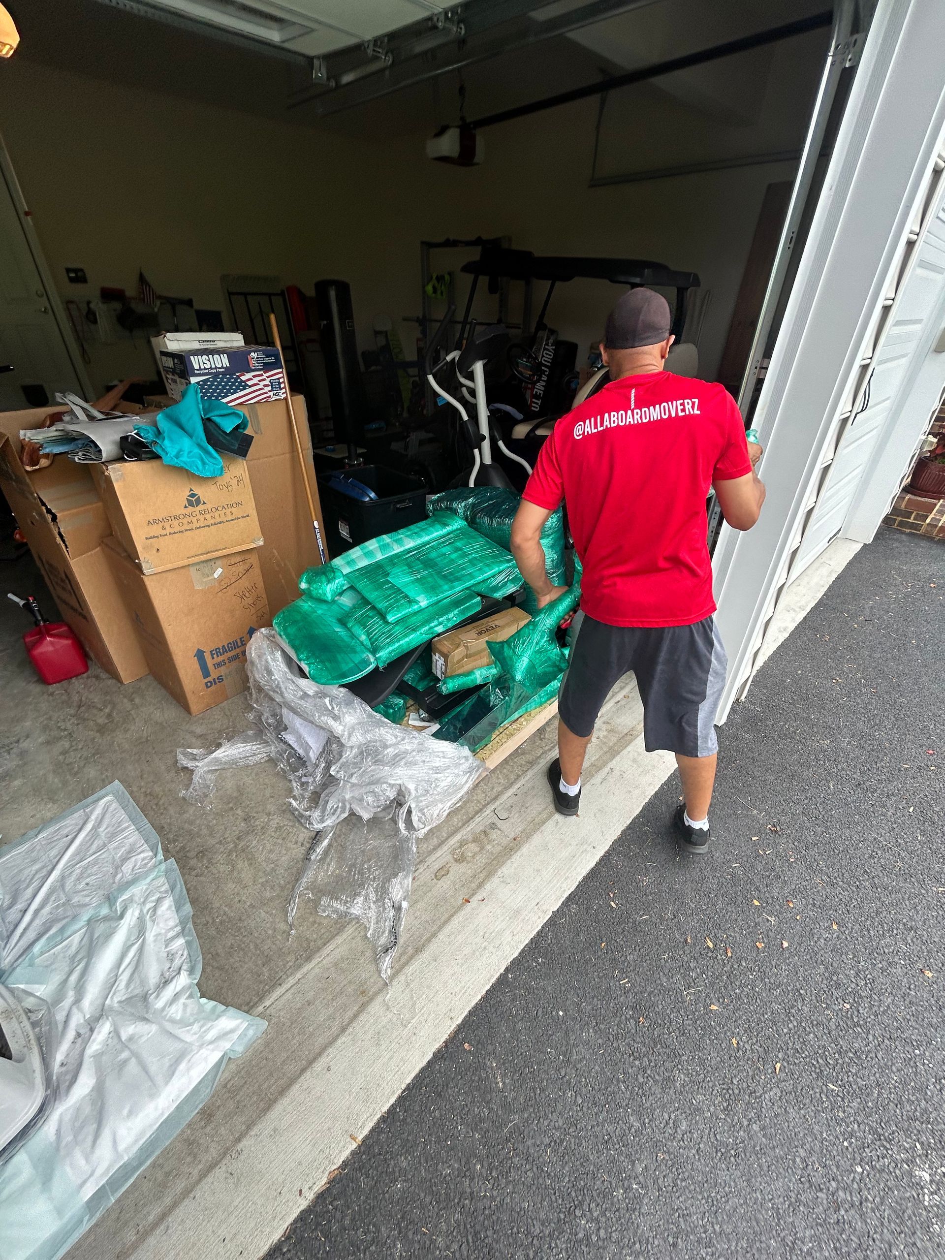 A man in a red shirt is carrying a green bag into a garage.