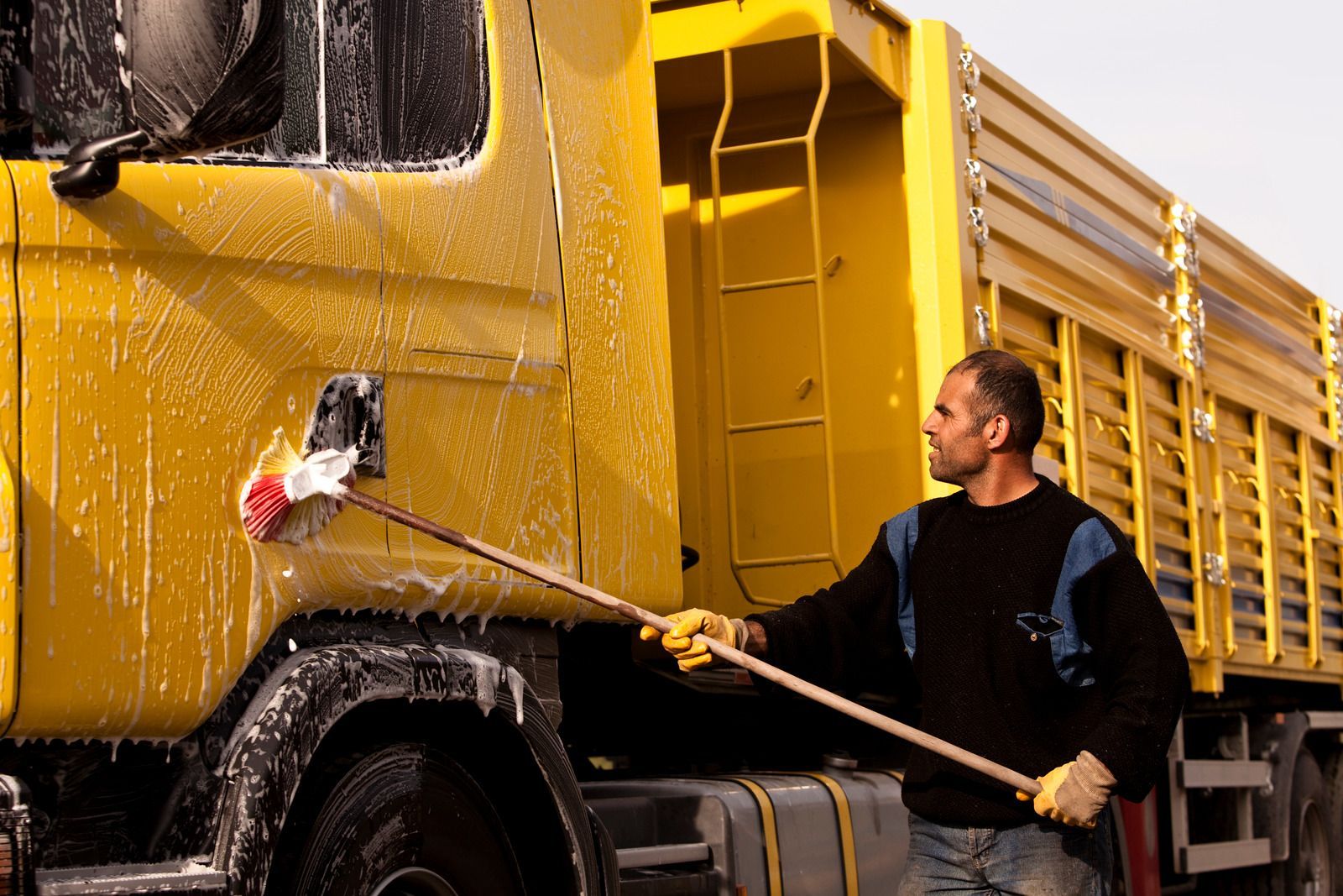 Man washing a yellow truck with a long-handled brush, outdoors.